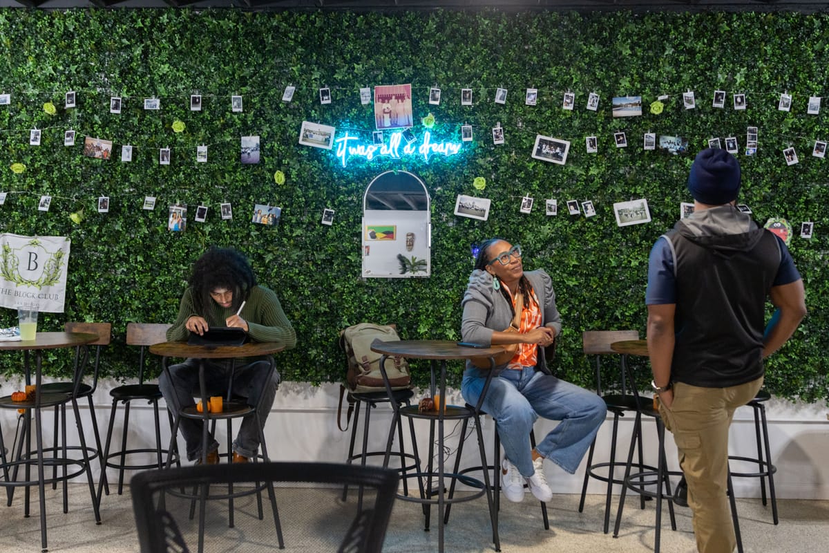 Two people sit at tables and one stand inside a venue in front of a wall covered in faux greenery and photographs