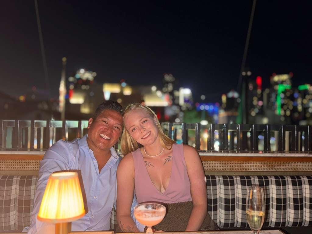 A man in a blue button-down shirt and a woman in a pink blouse sit at a table at a rooftop venue at nighttime