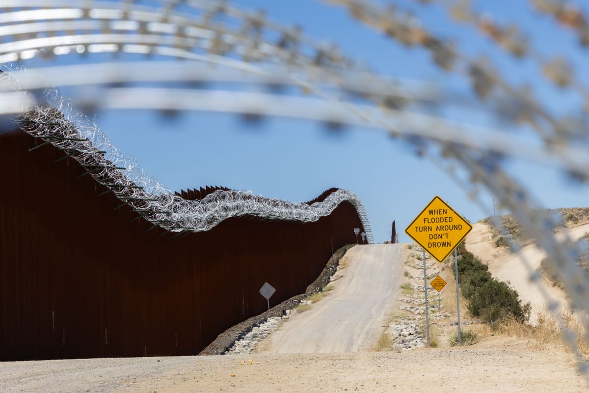 Concertina wire frames a view of a tall rust red border wall with concertina wire on top