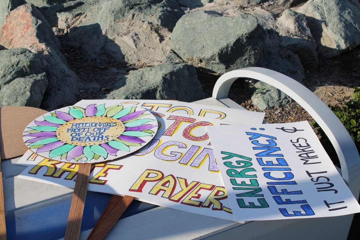 Handmade colorful signs are laid on a bench, reading "Billionaires profit off our deaths" and "Energy efficiency: it just makes cents."