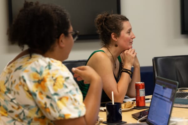 A woman in a floral dress with her back to the camera looks at a woman in a green dress who looks off to the right. Both sit at a table with laptops and food in front of them.