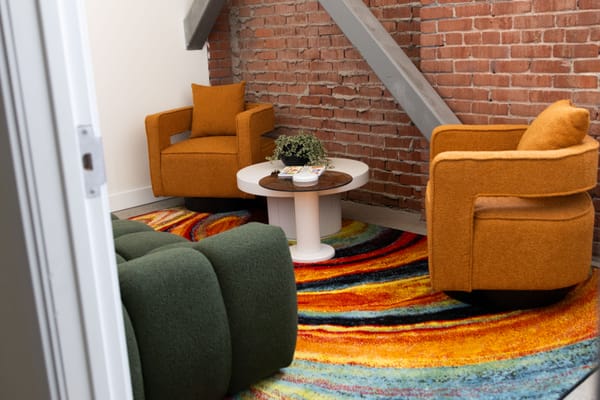 Two orange chairs and a green couch sit on a multi-colored rug in a small room with a white wall and a brick wall