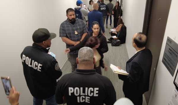 Five people stand around a woman looking backward in a hallway. More people stand around the background of the hallway as a person is walked away in handcuffs
