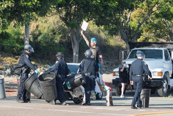 officers move trash cans out of the street as a woman with short, blue hair stands with a sign that says, "Melt ICE"