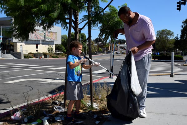 A man and his young son fill a trash bag while cleaning an area under a tree.