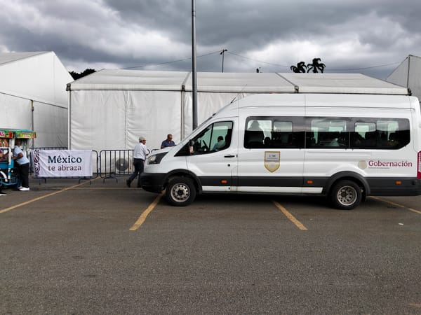 A white van with Gobernación written on the side is parked in front of a white tent with silhouettes of people leaving the van seen through the window