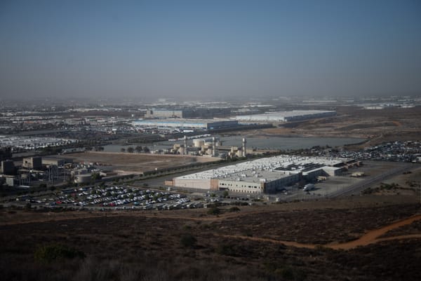 A prison-like building sits at the bottom of a hill with an Amazon warehouse in the background.