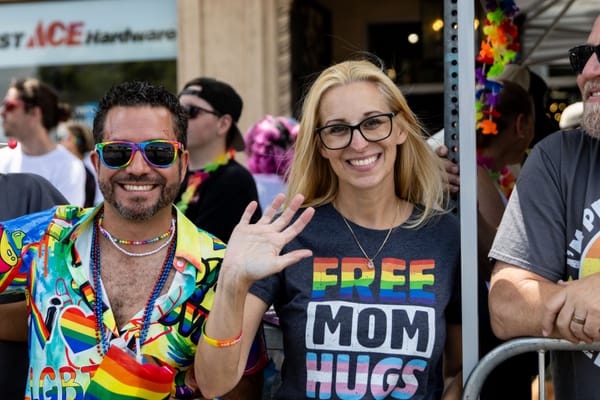 A person wearing a Hawaiian style shirt with LGBTQ+ logos on it and another person wearing a “Free Mom Hugs” shirt wave toward the camera.
