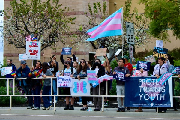 People stand outside a hospital at a protest holding signs that read "protect trans health" and "gender-affirming care saves lives"