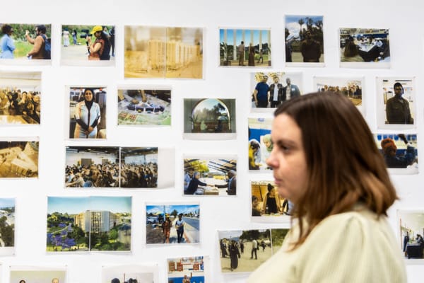 A woman with shoulder-length brown hair stands in front of a white wall covered in photos.