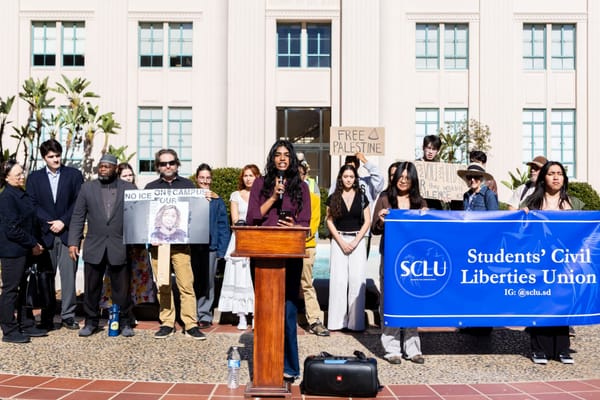 A crowd of people holding signs stand outside a building facing the camera while a woman speaks at the podium. 