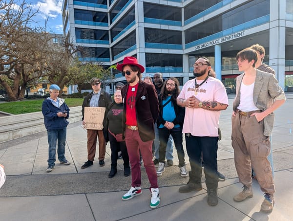 A man in red clothing and a hat stands at the front of a group of people in a plaza in front of a building with a sign that says San Diego Police Department