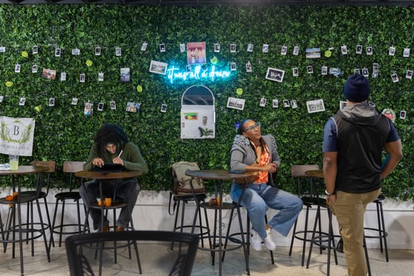 Two people sit at tables and one stand inside a venue in front of a wall covered in faux greenery and photographs