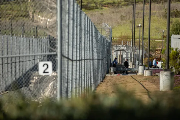 Two people walk out of a gate in a tall metal fence with razor wire on top