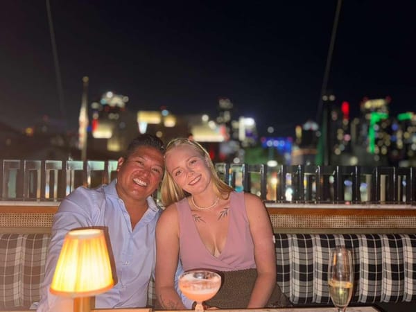 A man in a blue button-down shirt and a woman in a pink blouse sit at a table at a rooftop venue at nighttime