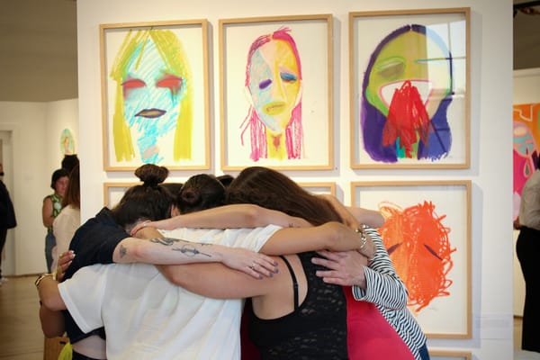 Women stand in a huddle with arms around one another and heads down, in front of art featuring many colorful painted faces.