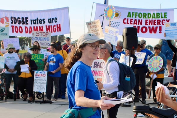 A woman with glasses in a cap and jacket tied around her waist holds paper and smiles in a group of protestors. 