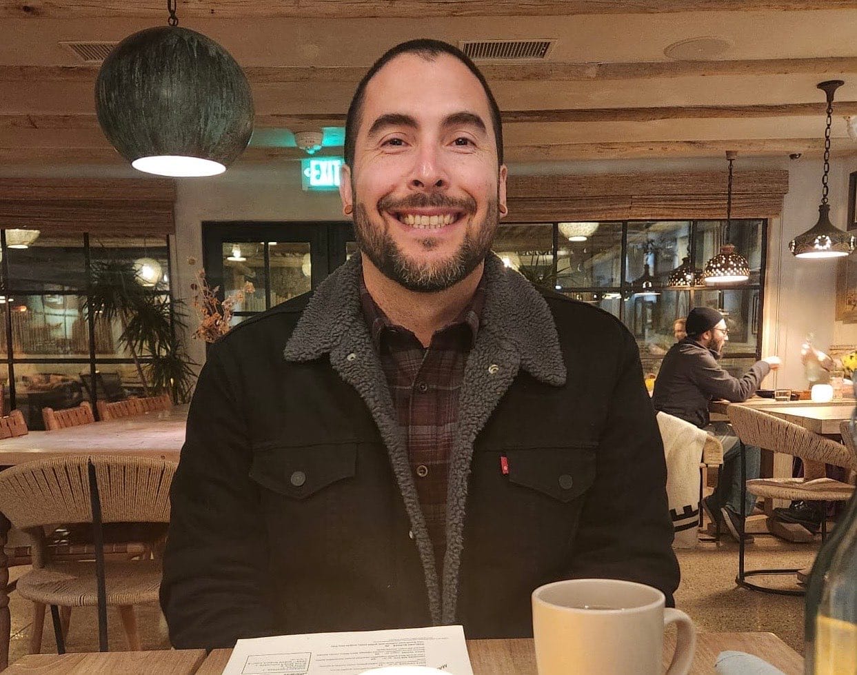 A man smiles sitting in a restaurant, wearing a black jacket